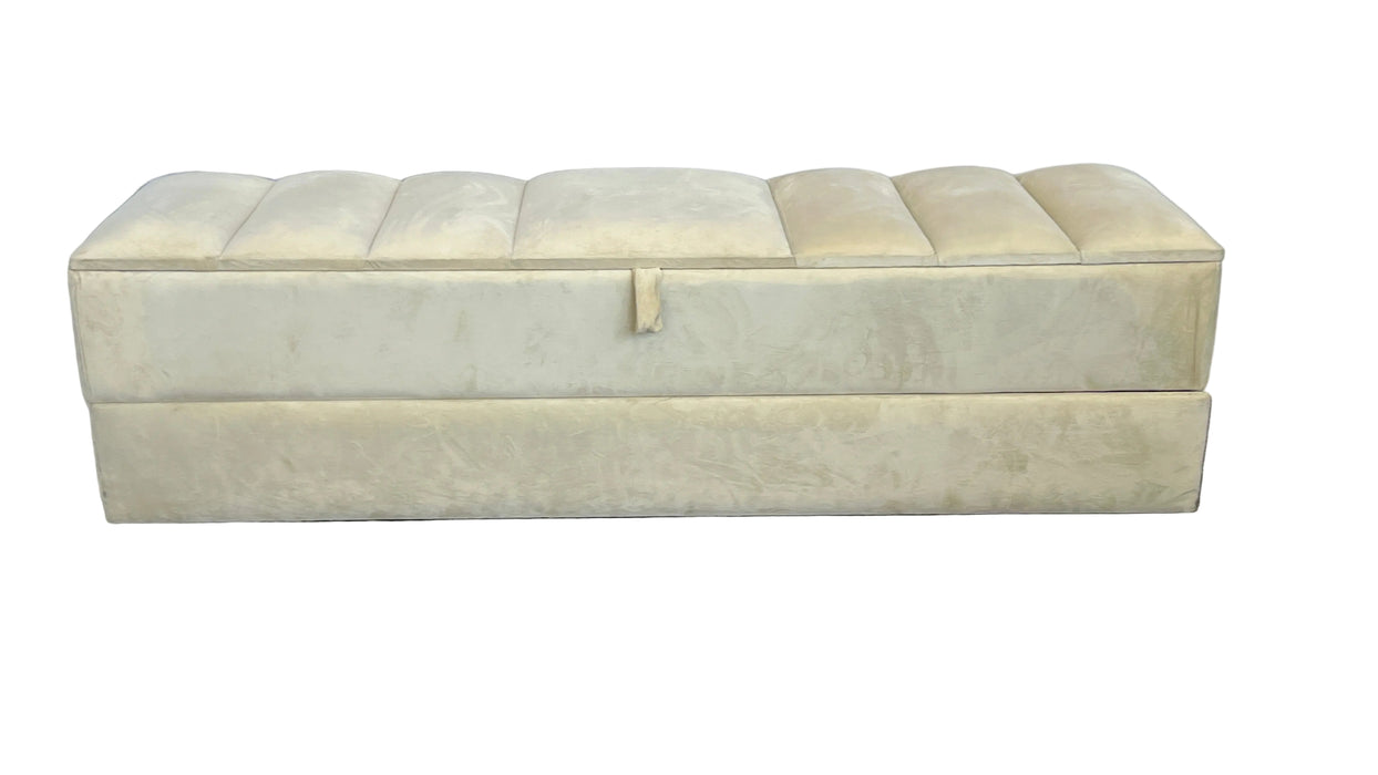 Beige upholstered storage bench on a carpeted floor with a white curtain background