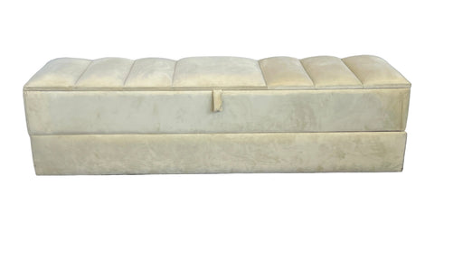 Beige upholstered storage bench on a carpeted floor with a white curtain background