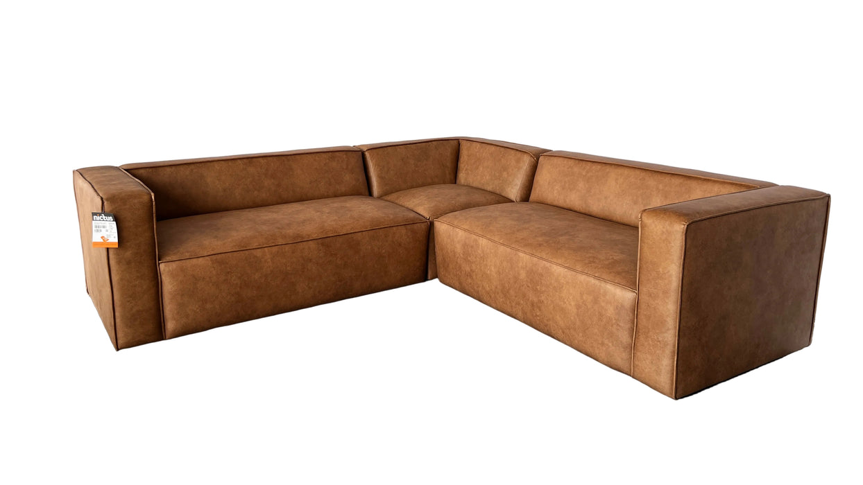 A modern L-shaped sectional sofa upholstered in medium brown textured faux leather, positioned on a gray floor in front of a white draped backdrop inside a showroom.