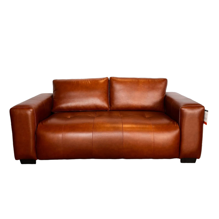 A brown leather loveseat sofa photographed from above on a white sheet, with a tiled wall and gray floor visible around it.